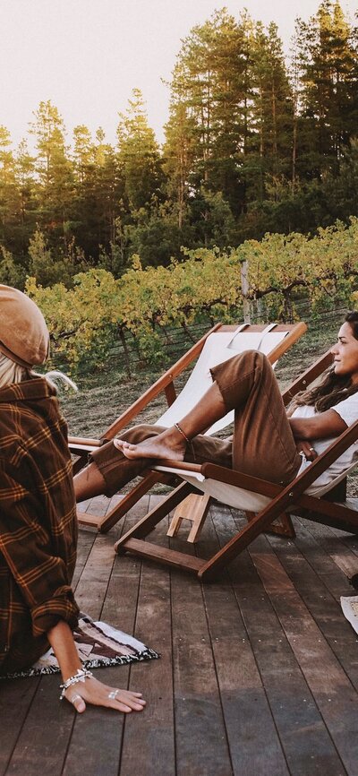 Two women drinking wine on a deck in front of a glamping tent with wine vines in the background.