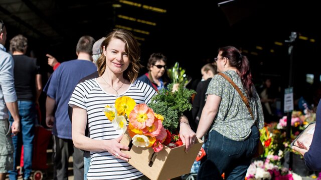 A woman holding a box full of fresh produce and poppies in front of the market.