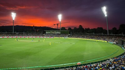 Cricketers playing on a green oval in front of a crowd at sunset.