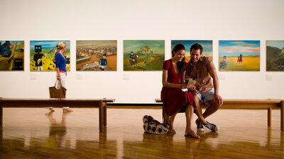 Two people sitting on a bench with art in the background.