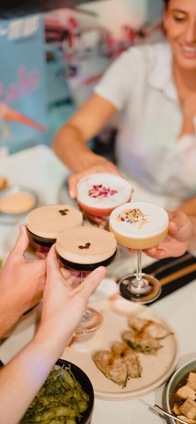 Two couples clinking cocktail glasses above a table laden with asian eats.