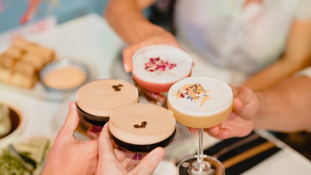 Two couples clinking cocktail glasses above a table laden with asian eats.