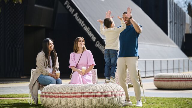 Two women sit outside and a man stands and high fives a young boy at the National Museum of Australia.