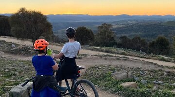 Two cyclists atop Mount Stromlo overlook an orange sunset.
