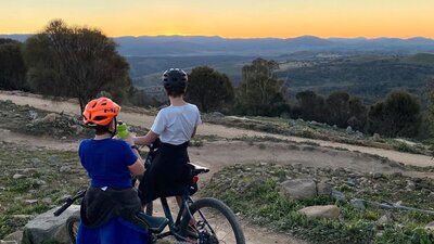 Two cyclists atop Mount Stromlo overlook an orange sunset.