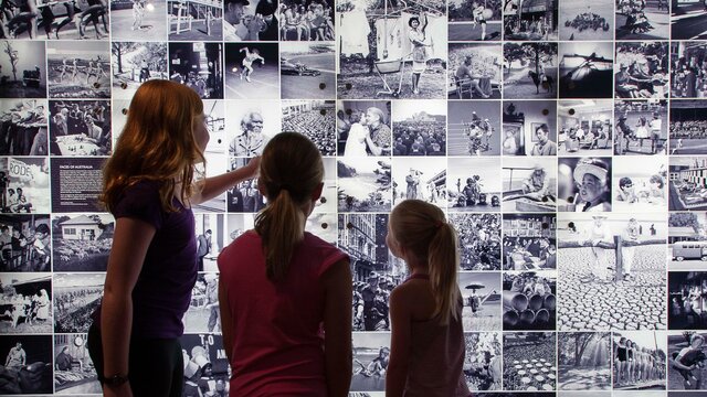 Three kids of varying heights looking at a digital collage of black and white photographs at the National Archives of Australia.