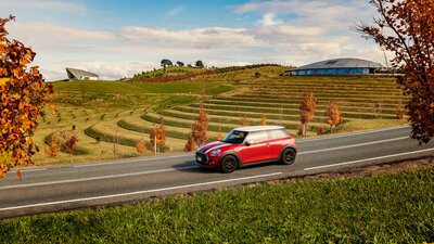 A red car driving on the road with autumn leaves near it and the National Arboretum Canberra in the background.